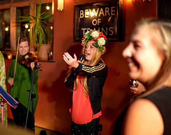 Ceilidh band caller instructs the dancers while wearing a floral crown, surrounded by musicians and smiling audience.