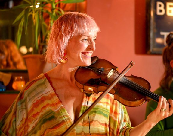 Smiling woman with pink hair playing a violin in a warm, vibrant setting.