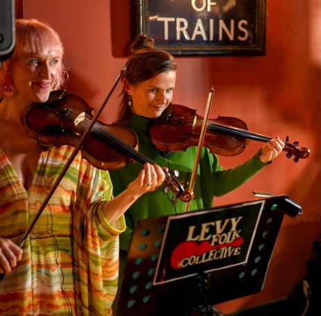 Two women playing violins in a lively indoor setting with a sign in the background.